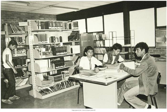  4 d- Fotografia do Colégio Agrícola, 1981 em que se retrata o Acervo do Arquivo Público/ DF. Um grupo de jovens está em uma biblioteca. Três desses jovens permanecem sentados em uma mesa clara, com alguns livros espalhados. Outro está próximo a duas prateleiras com diversos livros enfileirados. Realização: Academia Planaltinense de Letras, Artes e Ciências (APLAC), pelo Ecomuseu Pedra Fundamental e pelo Coletivo Nativo Audiodescrição produzida  pelo Instituto de Promoção das Pessoas com Deficiência Visual Audiodescritora: Elma Lúcia Rodrigues Consultor: Fernando Rodrigues Este projeto é promovido com recursos do Fundo de Apoio a Cultura do DF